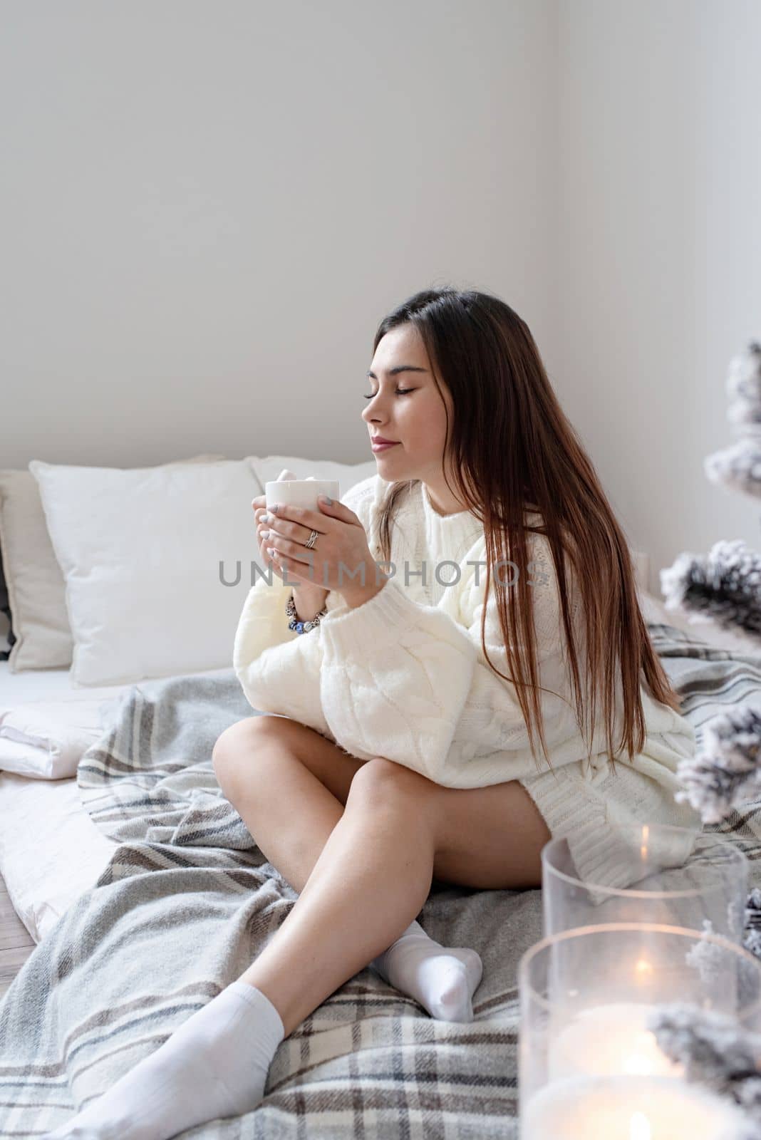 Merry Christmas and Happy New Year. Woman in warm white winter sweater lying in bed at home at christmas eve holding cup with marshmallows, fir tree behind