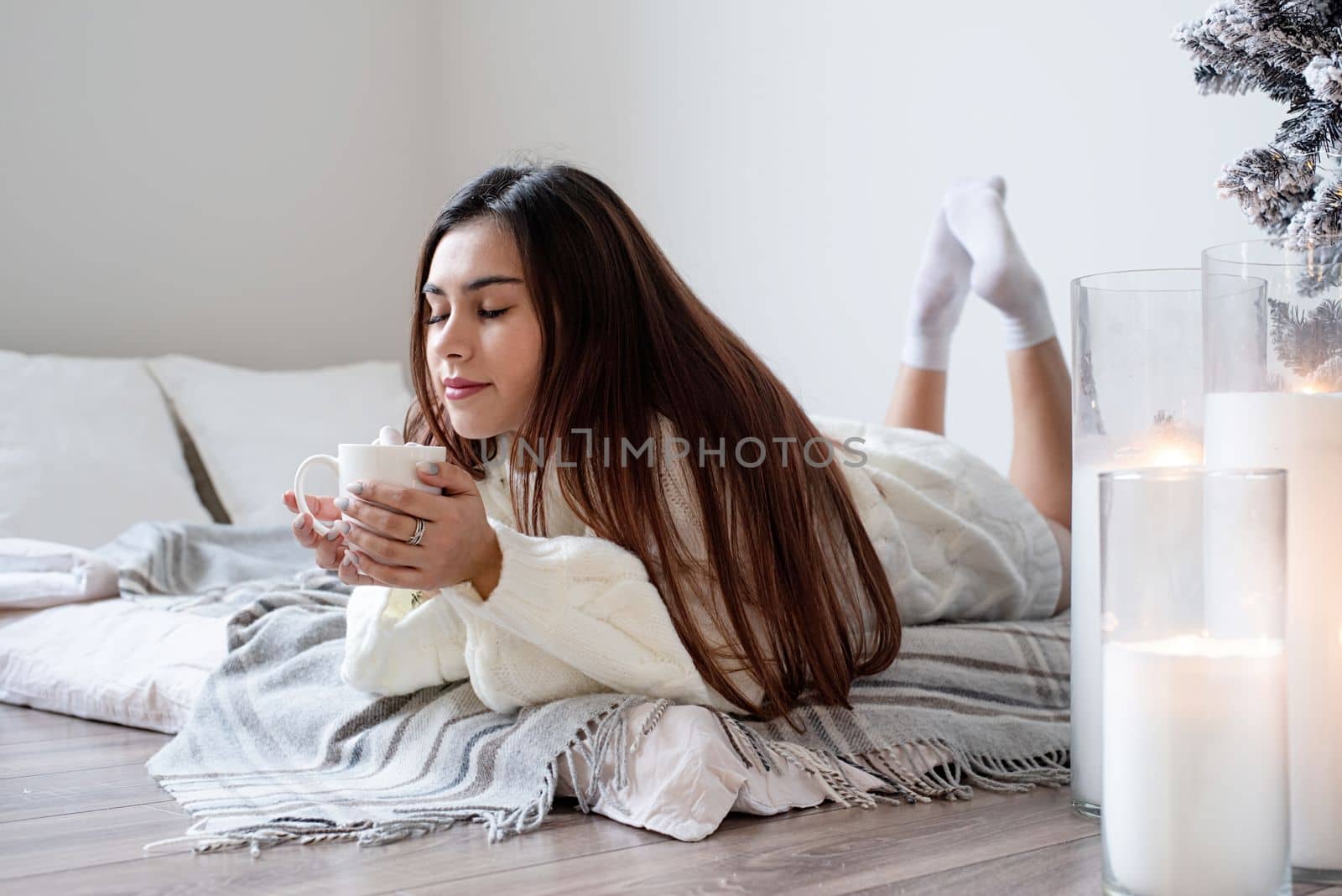 Woman in warm white winter sweater lying in bed at home at christmas eve holding cup with marshmallows, fir tree behind by Desperada