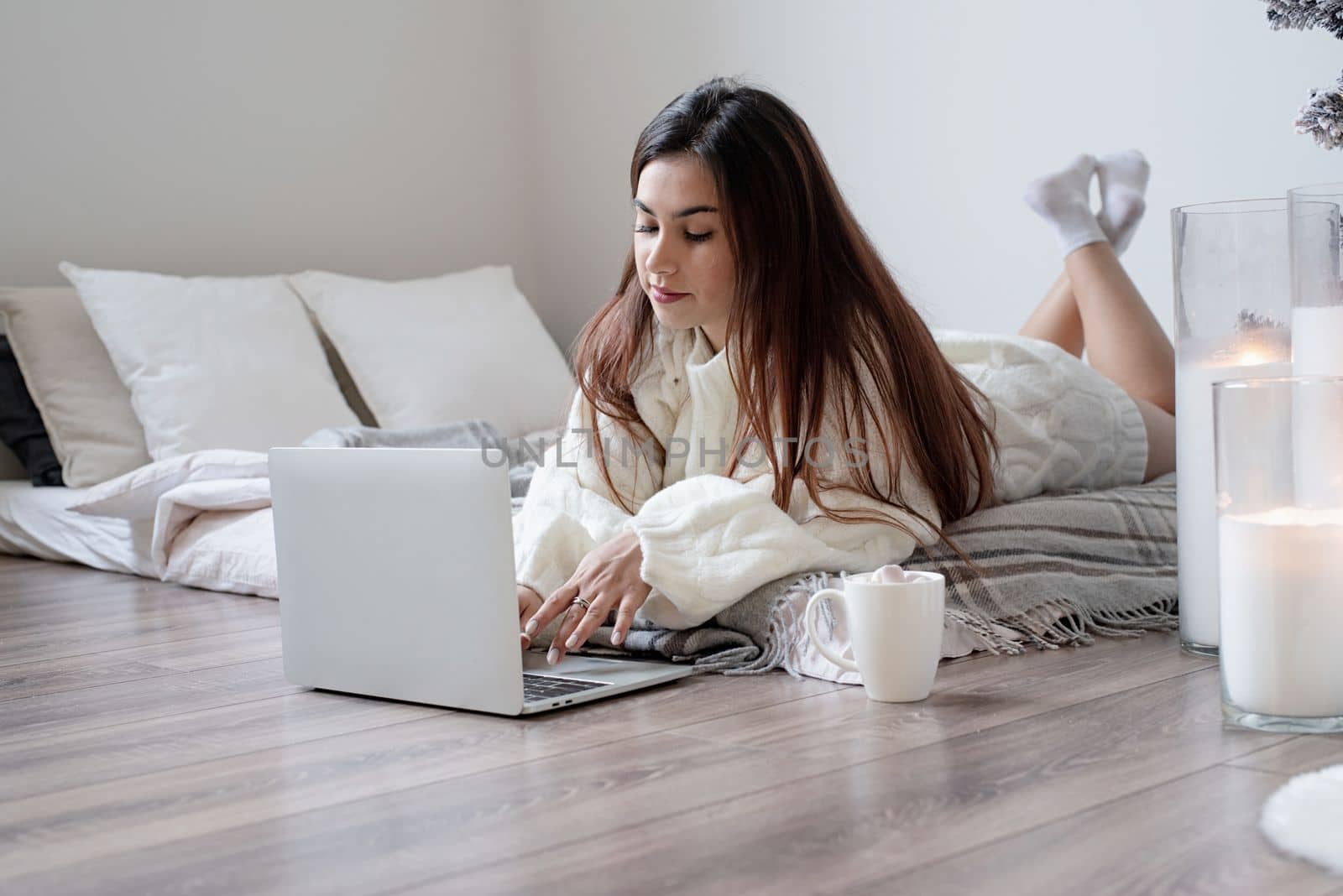 Merry Christmas and Happy New Year. Woman in warm white winter sweater lying in bed at home at christmas eve holding cup with marshmallows, fir tree behind