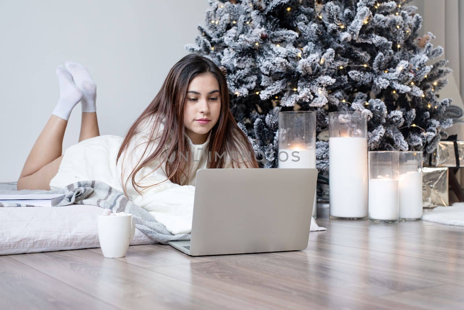 Woman in warm white winter sweater lying in bed at home at christmas eve holding cup with marshmallows, fir tree behind by Desperada