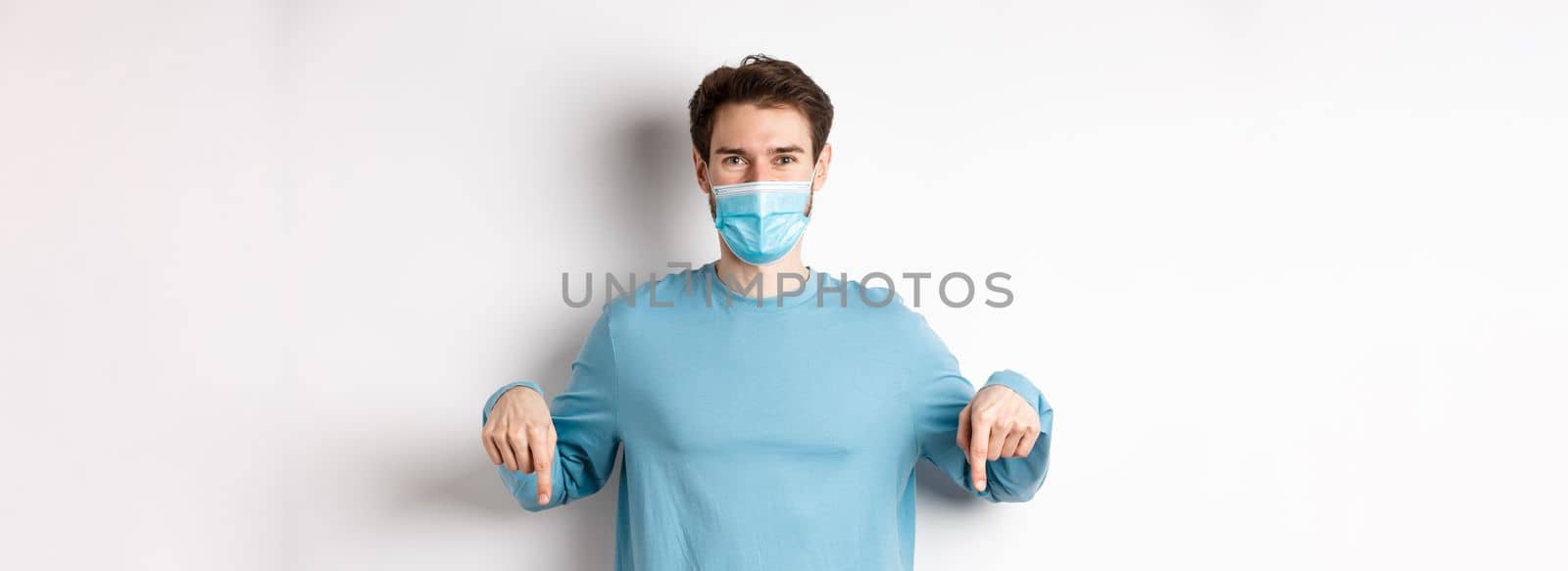 Covid-19, health and quarantine concept. Smiling caucasian man in face mask pointing fingers down, showing logo, standing over white background by Benzoix