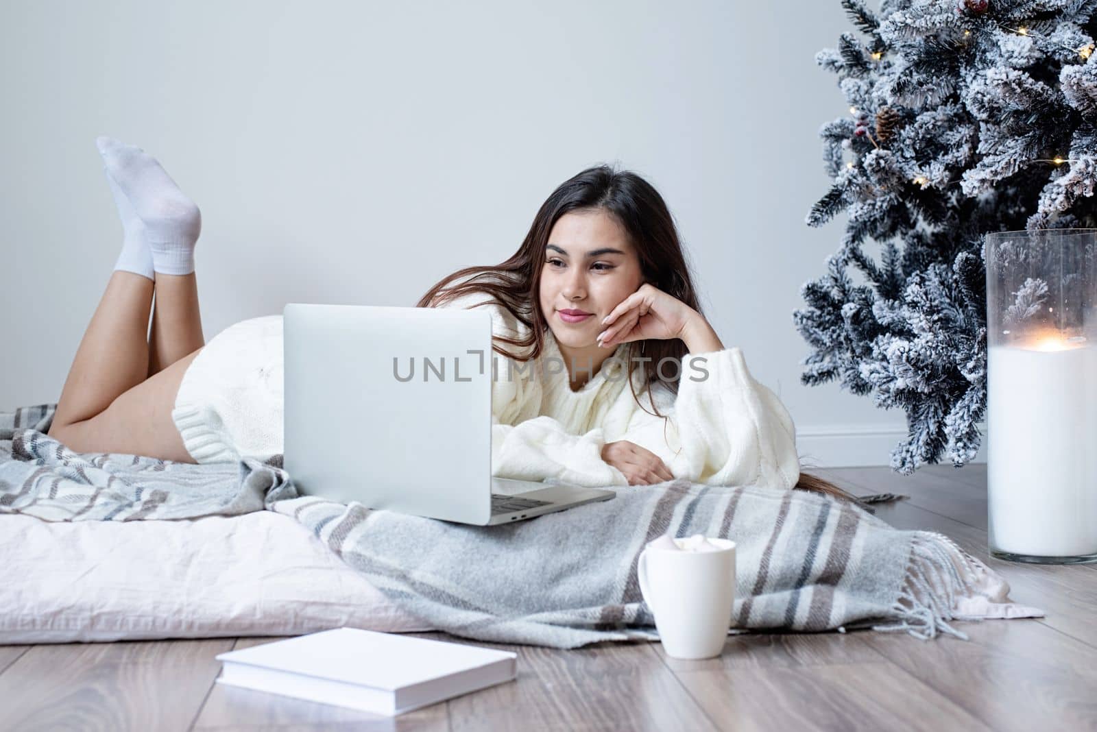 Merry Christmas and Happy New Year. Woman in warm white winter sweater lying in bed at home at christmas eve holding cup with marshmallows, fir tree behind