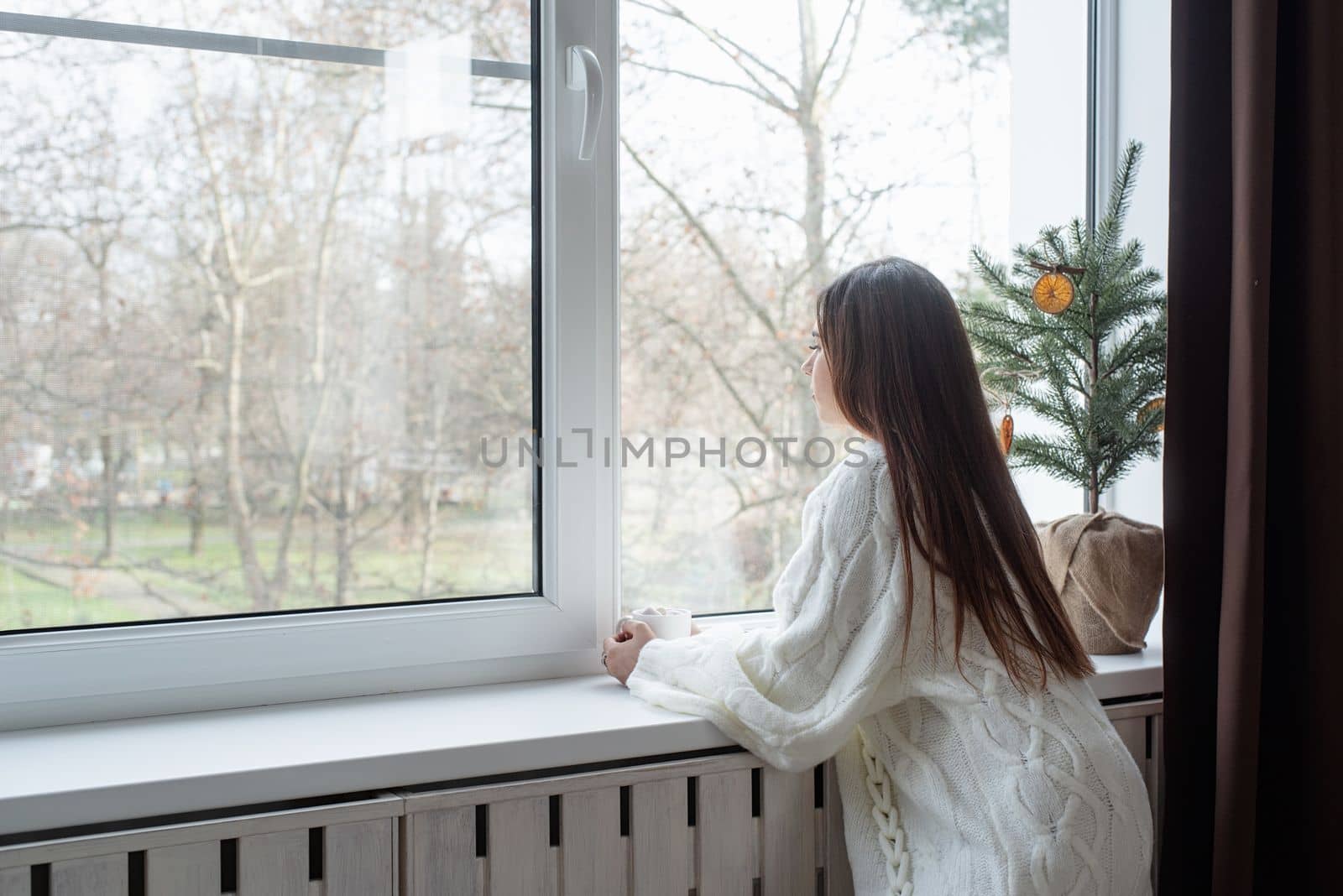 Merry Christmas and Happy New Year. Woman in warm white winter sweater standing next to the window at home at christmas eve holding cup with marshmallows, fir tree behind