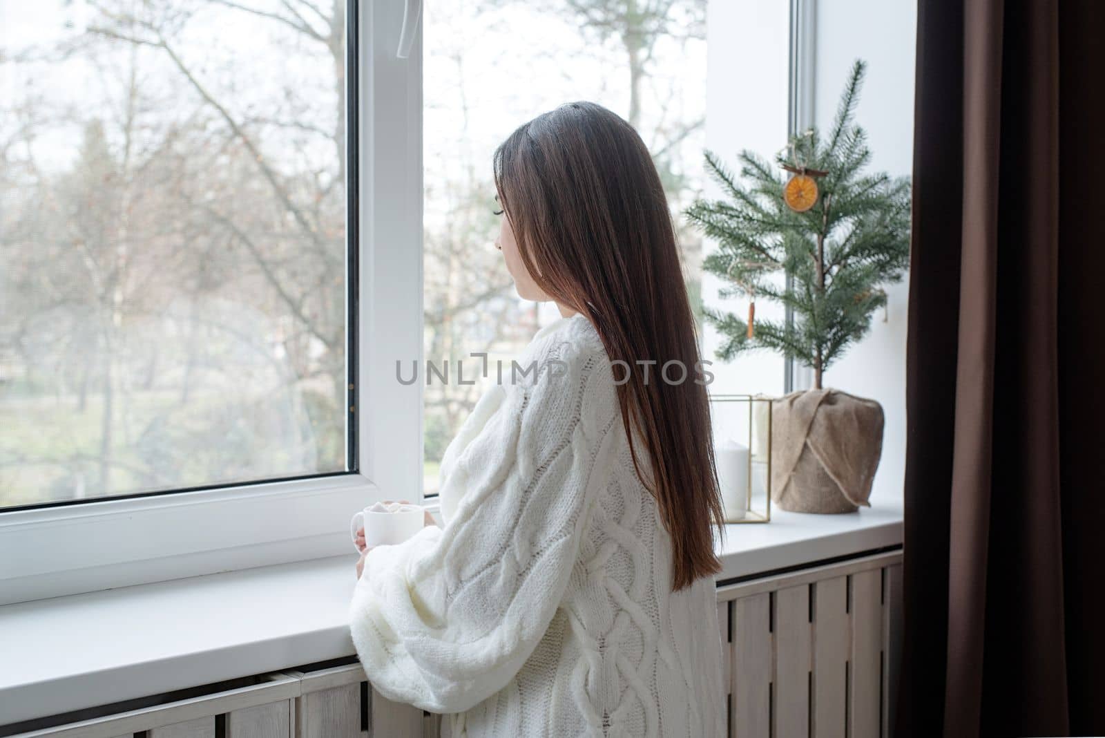 Merry Christmas and Happy New Year. Woman in warm white winter sweater standing next to the window at home at christmas eve holding cup with marshmallows, fir tree behind