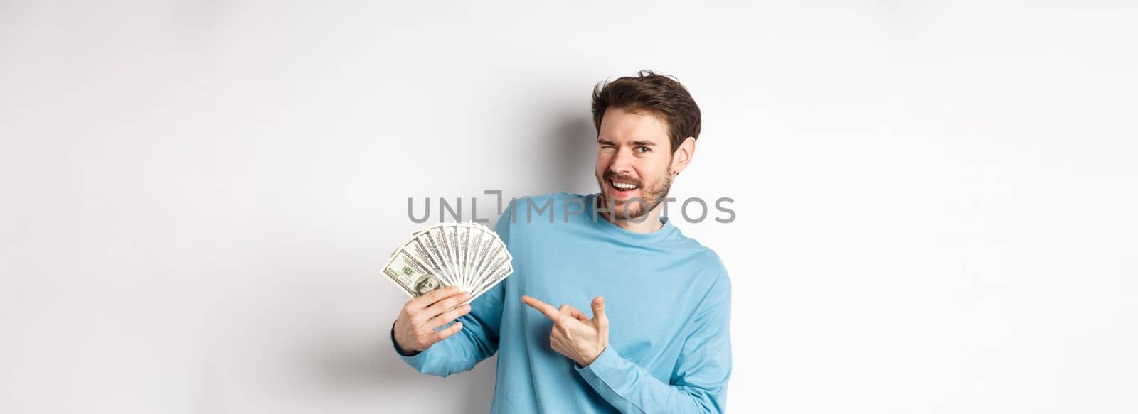 Handsome rich guy winking at camera, showing income, pointing finger at earned money and smiling, standing over white background by Benzoix