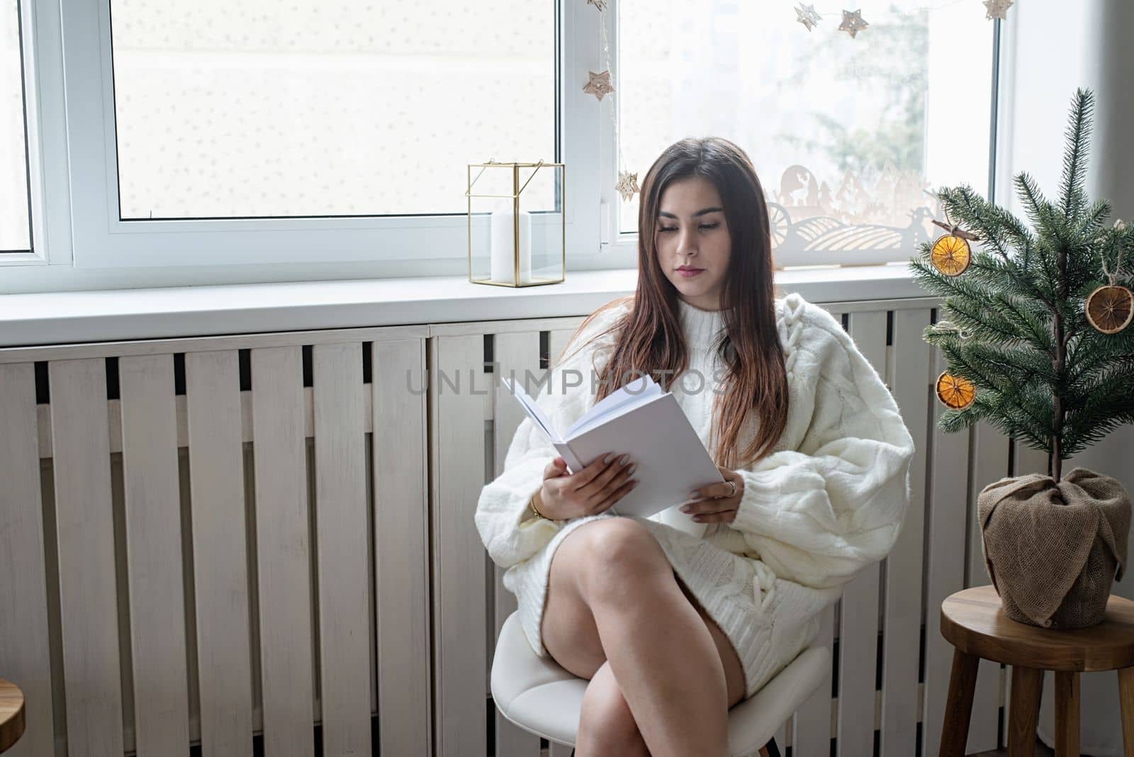 Merry Christmas and Happy New Year. Woman in warm white winter sweater lying in bed at home at christmas eve holding cup with marshmallows, fir tree behind