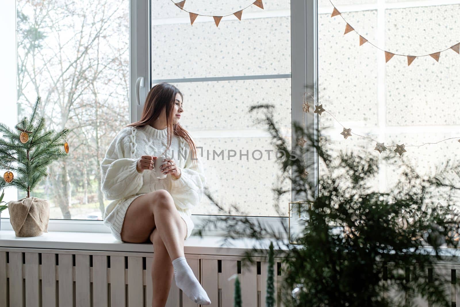 Merry Christmas and Happy New Year. Woman in warm white winter sweater sitting on the window at home at christmas eve holding cup with marshmallows, fir tree behind