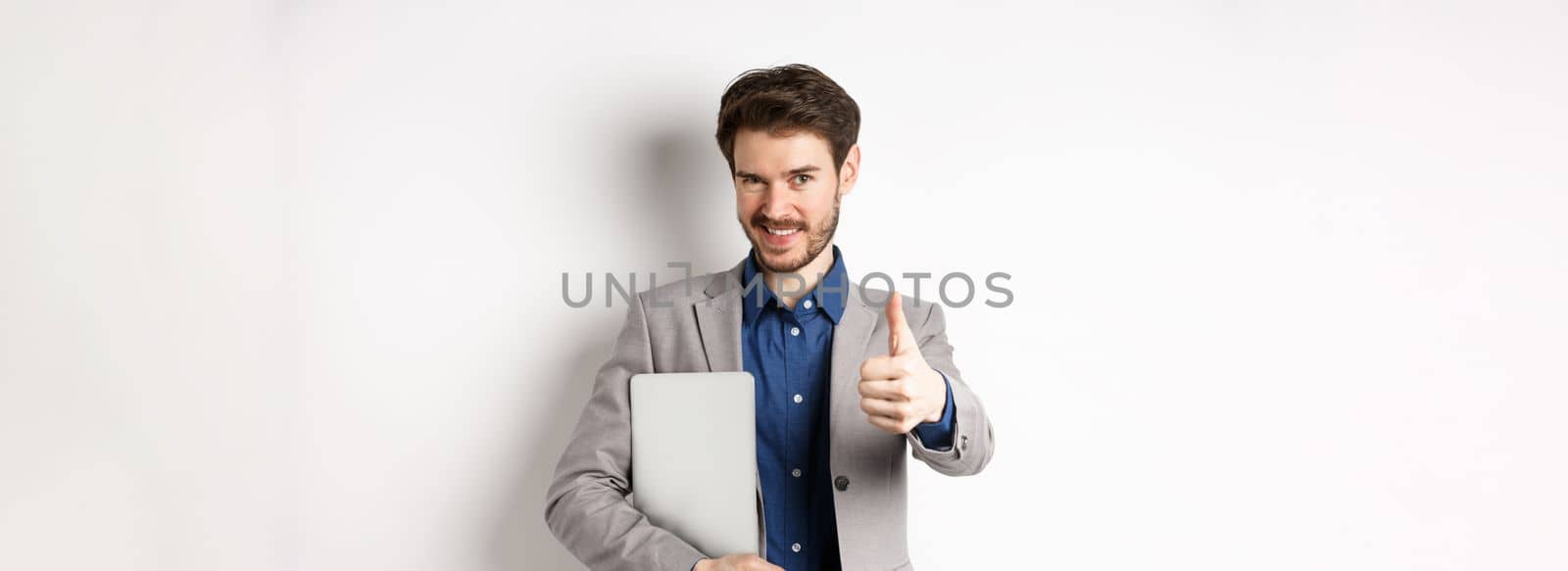 Successful businessman in stylish suit carry laptop and looking confident at camera, showing thumbs up in approval, white background by Benzoix