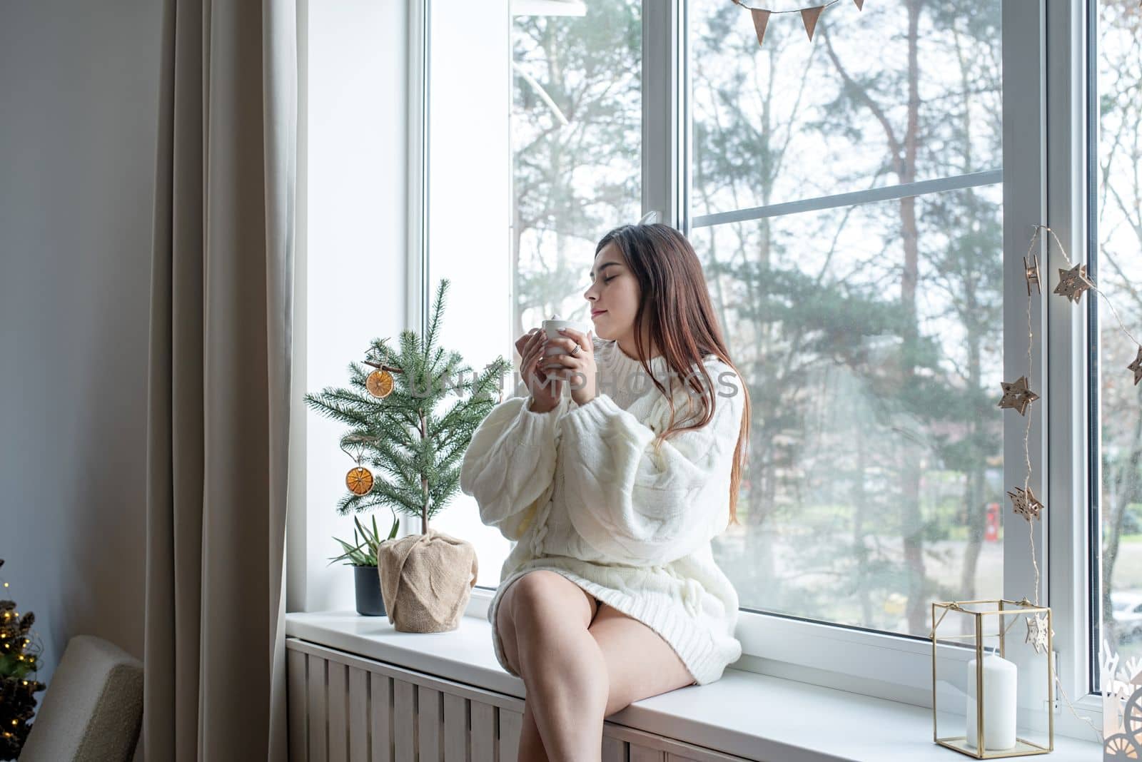 Merry Christmas and Happy New Year. Woman in warm white winter sweater lying in bed at home at christmas eve holding cup with marshmallows, fir tree behind