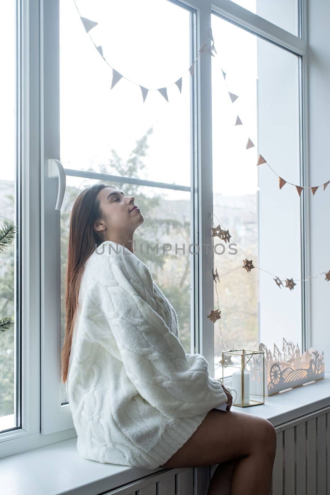 Woman in warm white winter sweater sitting at windowsill at home at christmas eve holding cup with marshmallows by Desperada