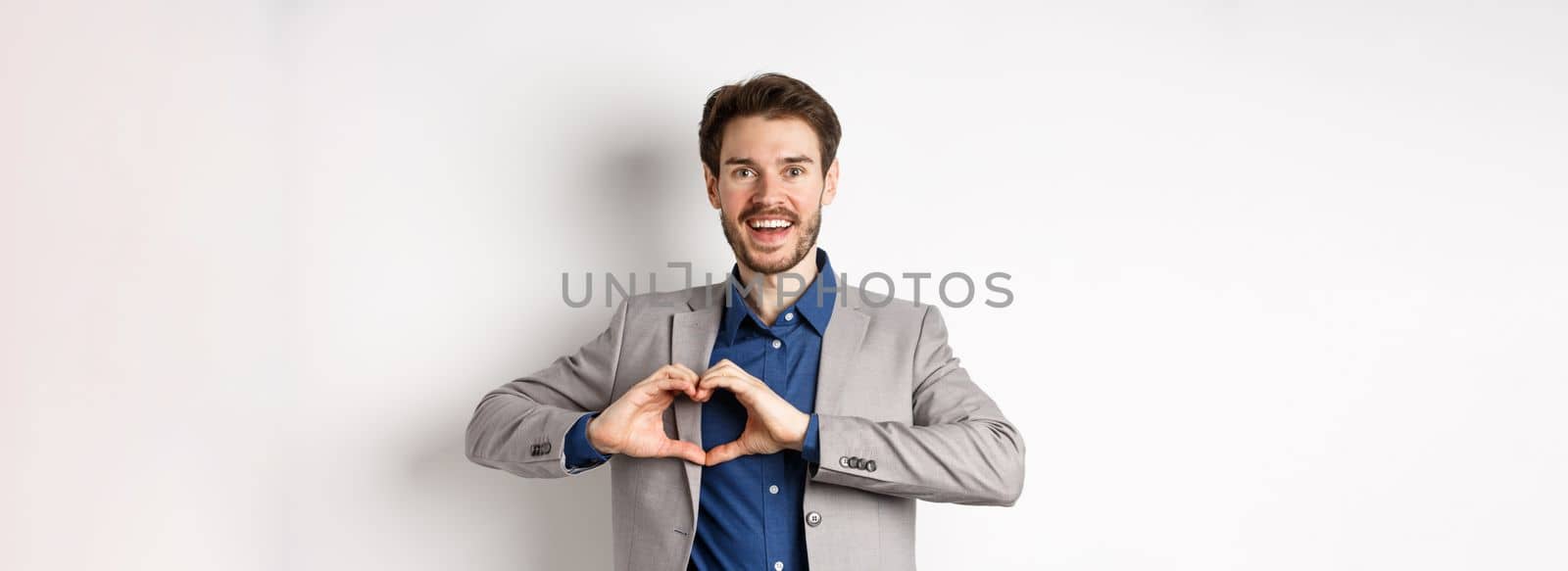 Passionate and romantic man in suit falling in love, showing heart sign and smiling at lover, standing on white background by Benzoix