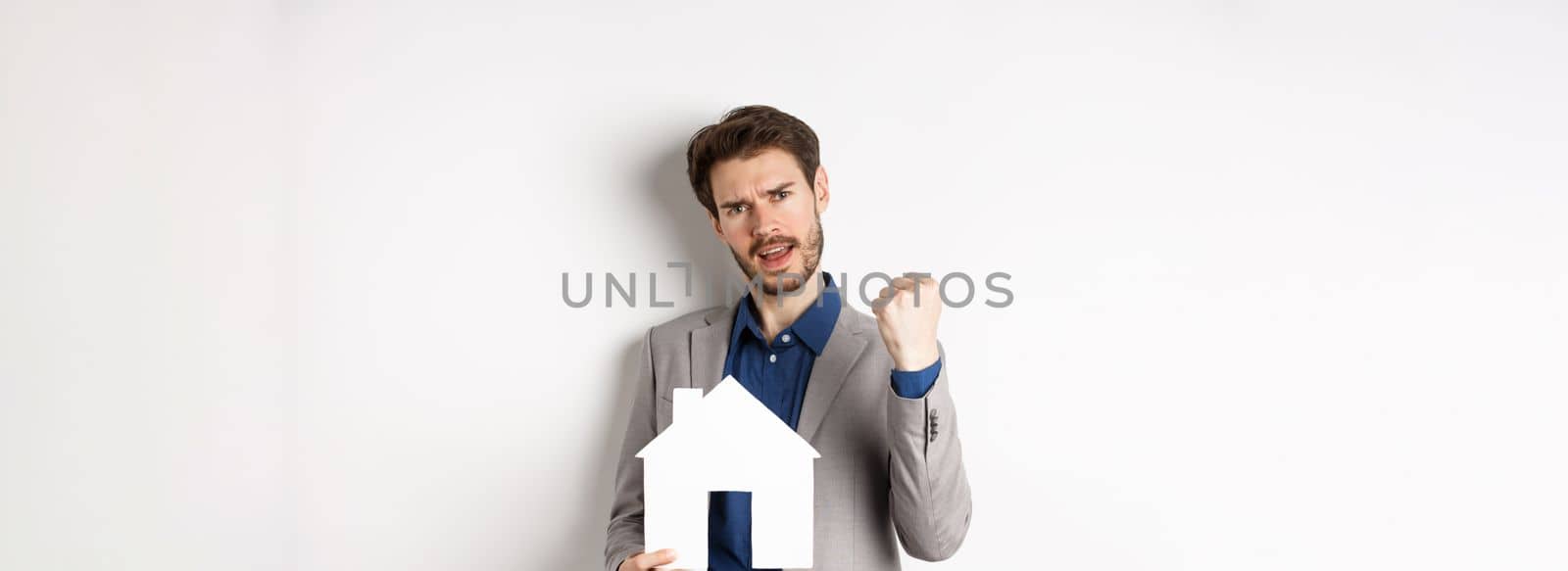 Real estate. Excited man buying house and showing paper home cutout, say yes and fist pump with joy, standing in suit on white background.