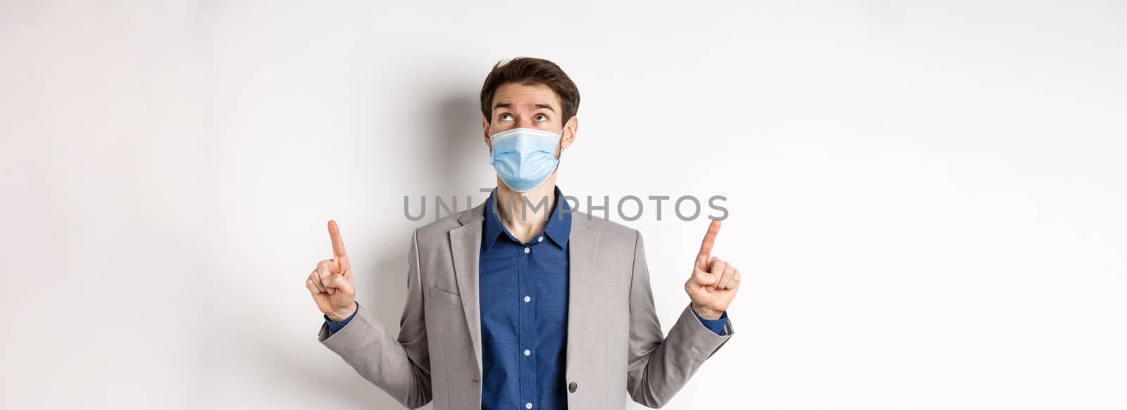 Covid-19, pandemic and business concept. Handsome businessman in medical mask and suit, looking and pointing up with dreamy face, white background.