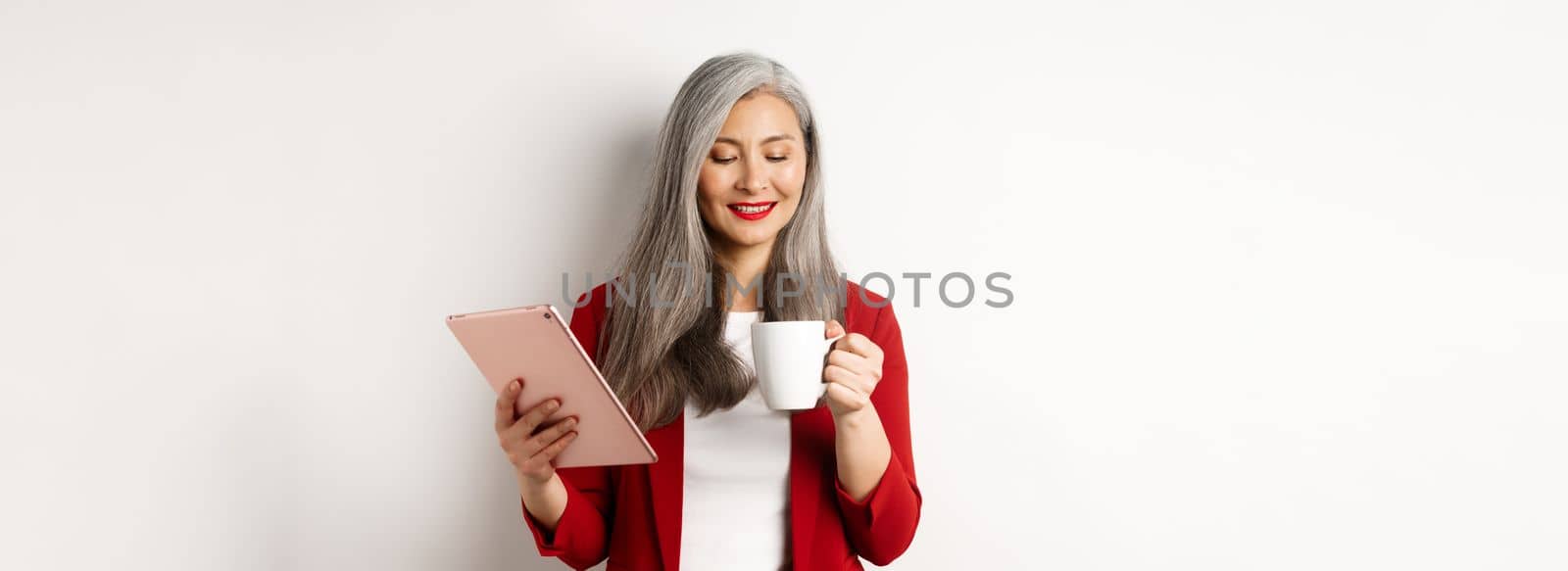 Business people concept. Asian mature businesswoman holding digital tablet and looking at coffee cup with pleased smile, standing over white background.