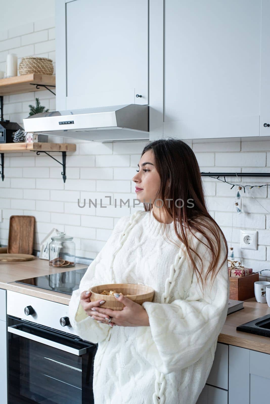 Merry Christmas and Happy New Year. Woman in warm white winter sweater standing at the kitchen at home holding bowl with popcorn