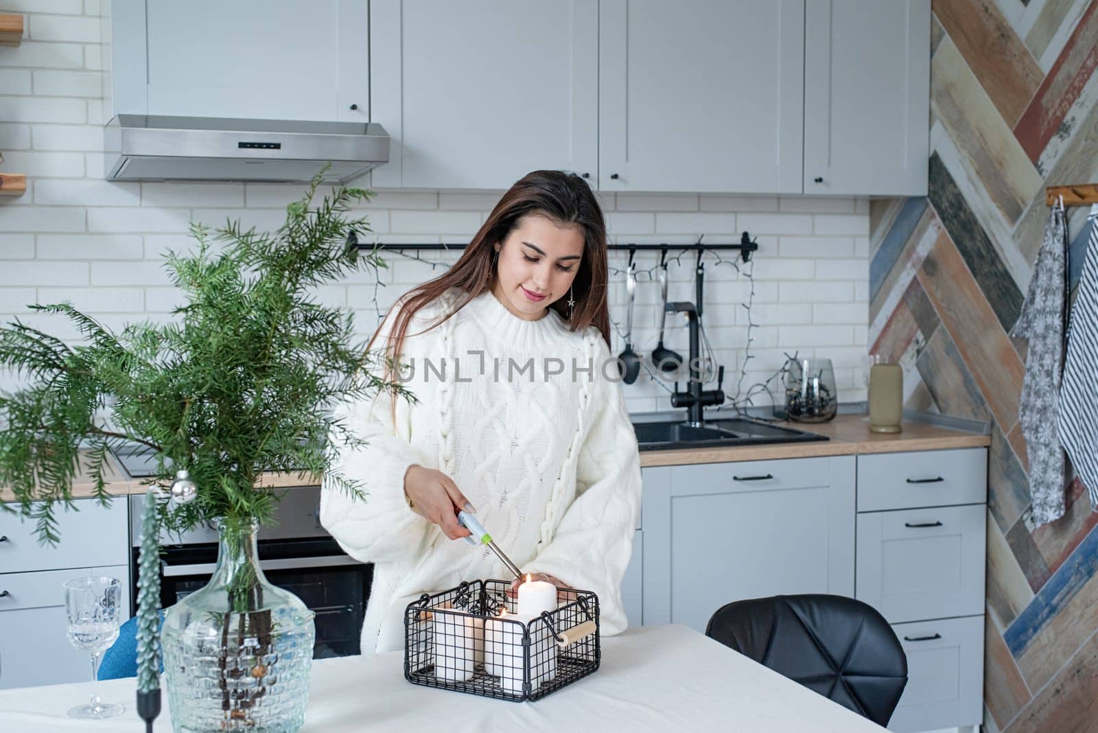 Woman in warm white winter sweater standing at the kitchen at home lighting up the candles by Desperada