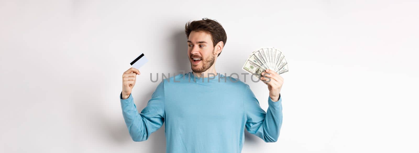 Handsome bearded man choosing between money and plastic credit card, payment with cash or contactless, standing over white background.