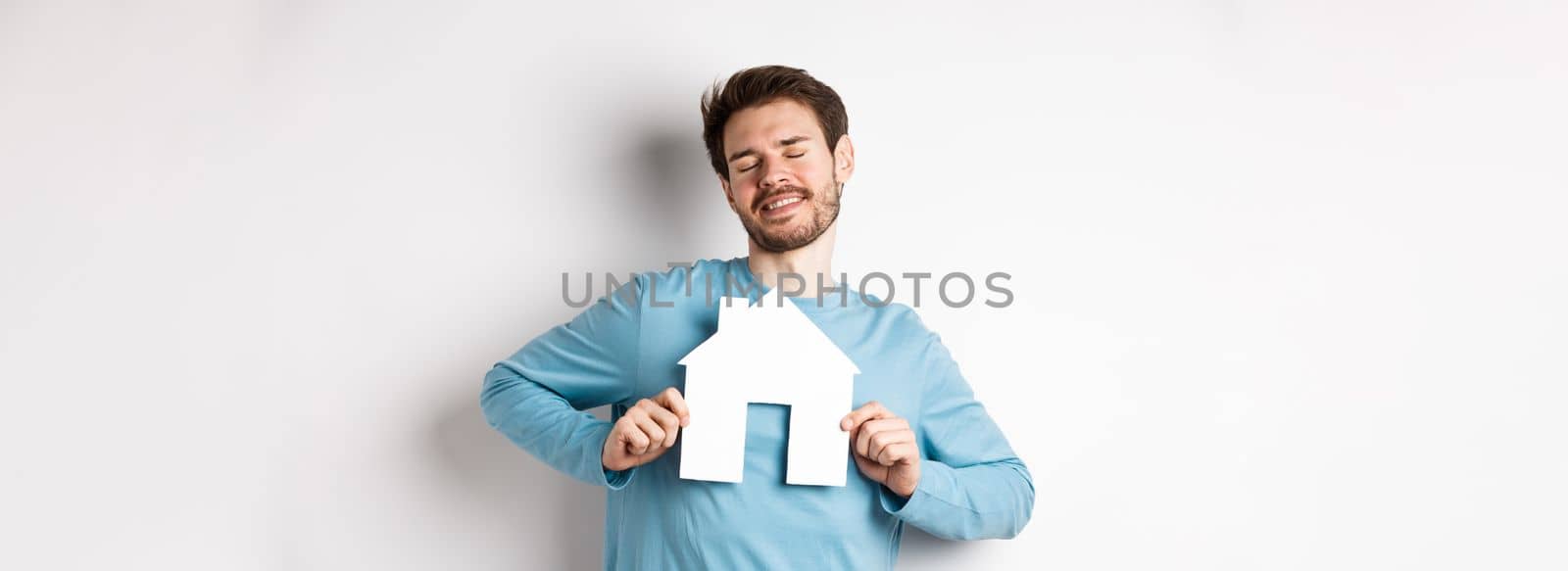 Real estate and insurance concept. Dreamy young man smiling with closed eyes, showing paper house cutout, wishing to buy home, standing over white background.