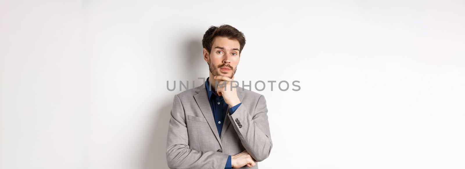 Thoughtful businessman in suit touching beard, thinking and looking at camera, deciding with pensive face, standing on white background.