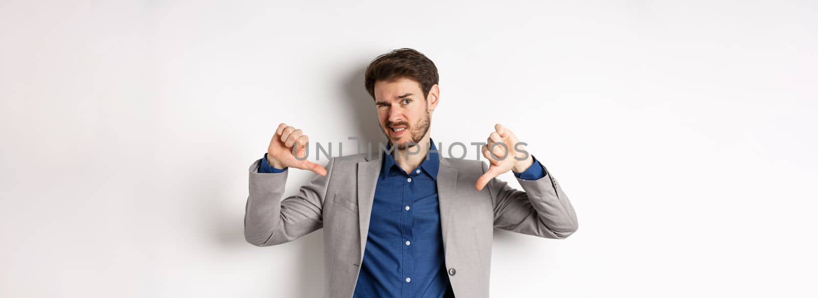 It sucks. Man feeling awkward showing thumbs down and judging something bad, frowning disappointed, standing in suit on white background.