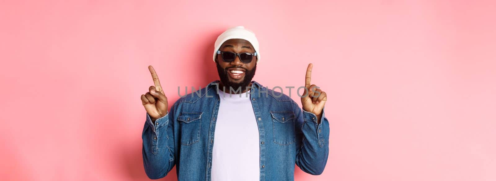 Happy bearded african-american man showing announcement, pointing fingers up and smiling, standing in beanie and sunglasses over pink background.