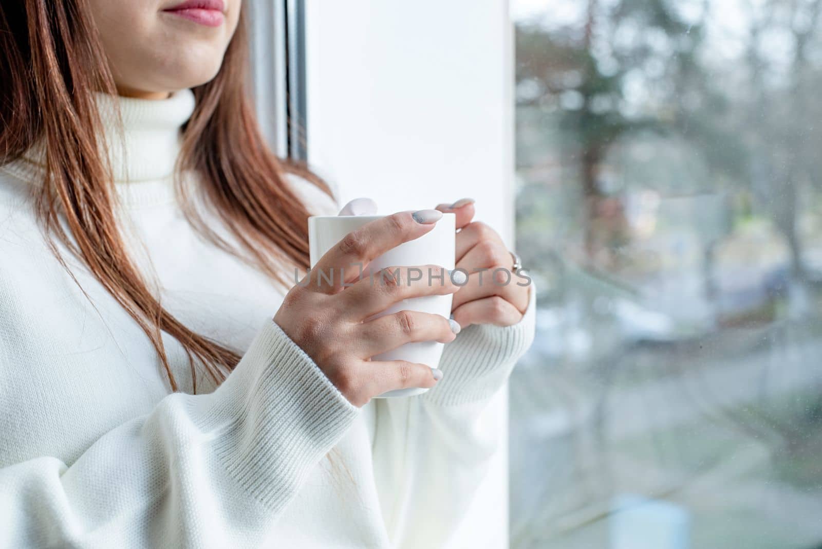 Merry Christmas and Happy New Year.Woman in warm white winter sweater sitting at windowsill at home at christmas eve working on laptop, fir tree behind