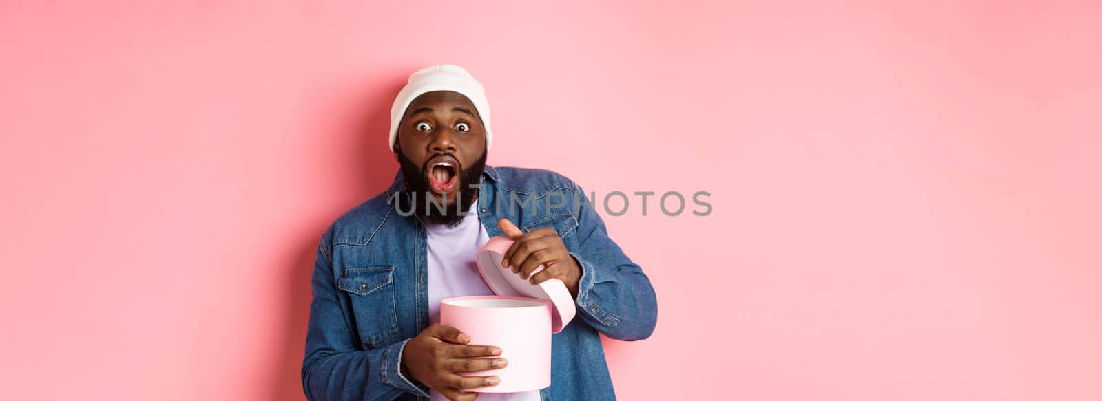 Excited Black man open box with gift and staring at camera grateful and amazed, receiving present, standing over pink background.