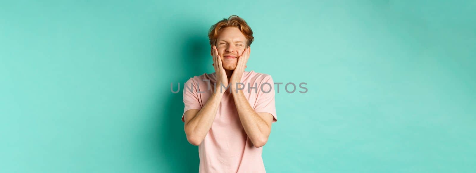 Image of cute and silly young man with red hair, feeling satisfaction while touching his face, smiling happy, standing over turquoise background.