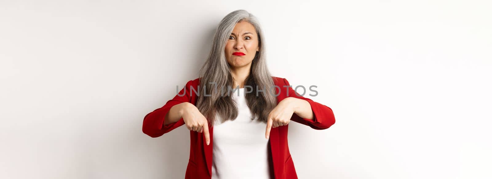 Disappointed and angry asian employer pointing fingers down, looking displeased and upset, standing over white background.