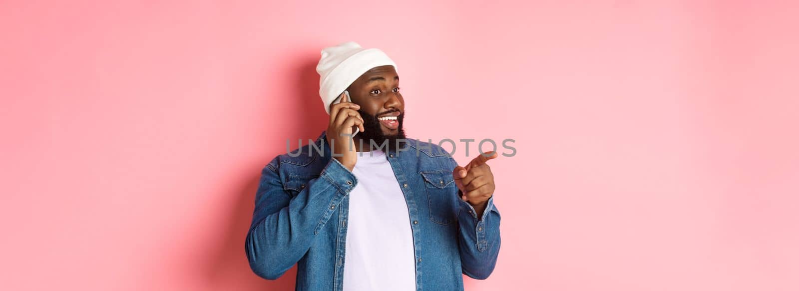 Handsome modern Black man talking on mobile phone, pointing left at person and smiling, standing over pink background.