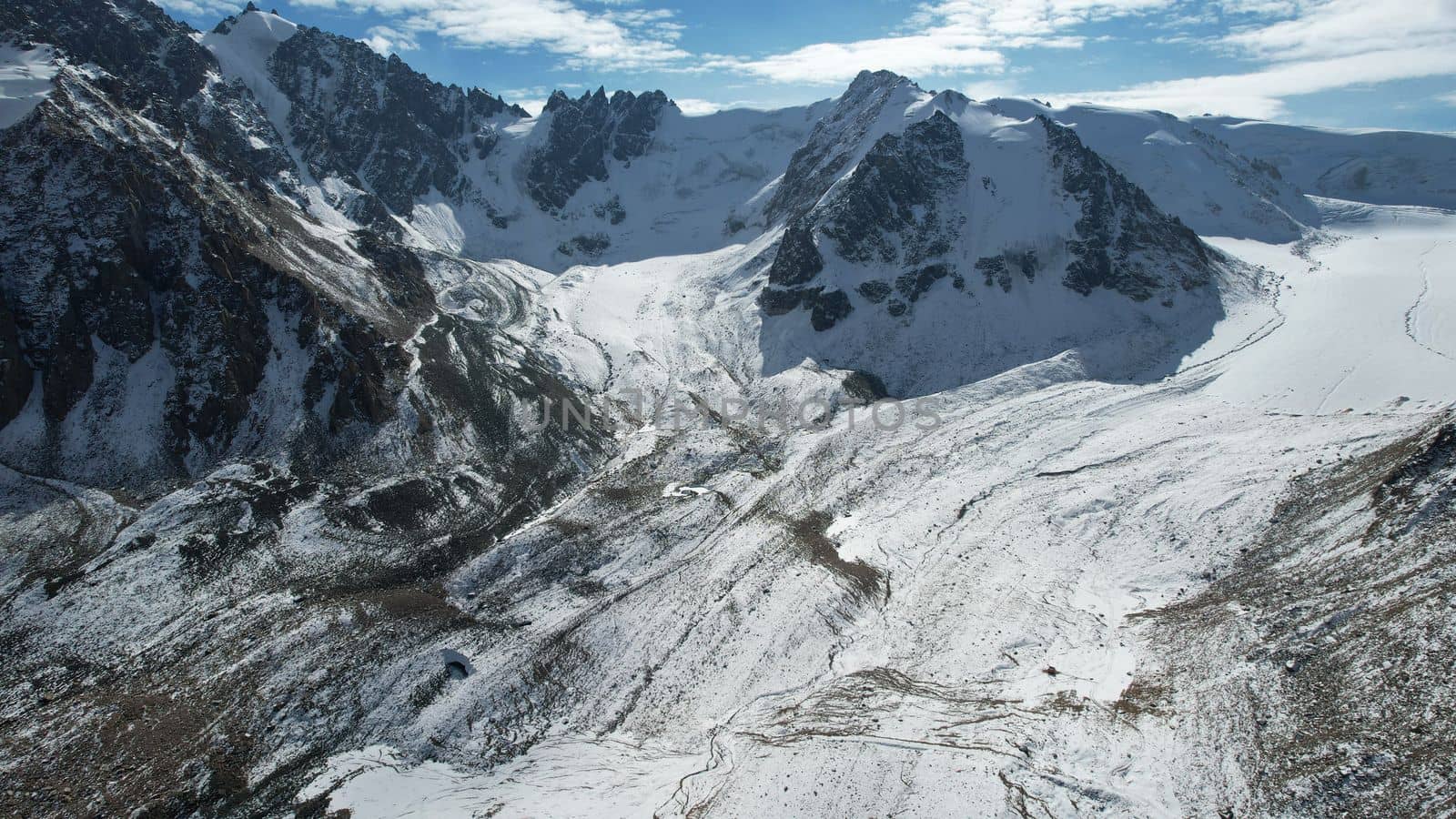 An ancient snow glacier among high mountains. The peaks of the mountains are covered with snow. In places there are steep cliffs and large rocks. Blue sky with white clouds. Top view from a drone