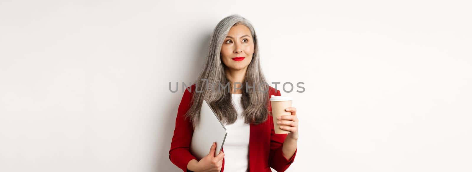 Business. Successful asian businesswoman with grey hair, drinking coffee and standing with laptop, looking thoughtful upper left corner.