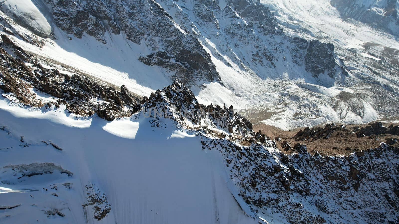 An ancient snow glacier among high mountains. The peaks of the mountains are covered with snow. In places there are steep cliffs and large rocks. Blue sky with white clouds. Top view from a drone