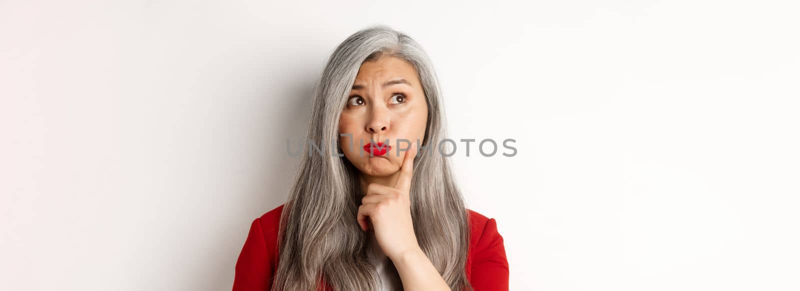 Close up of indecisive asian senior woman making choice, pouting and looking thoughtful at upper left corner, standing against white background by Benzoix