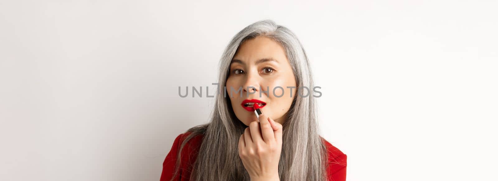 Beauty and makeup concept. Stylish asian mature woman with grey hair, looking in mirror and apply red lipstick, standing over white background.
