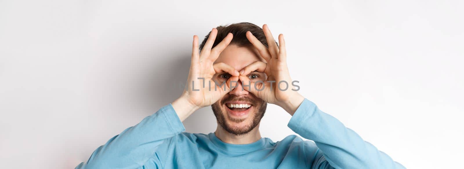 Happy smiling man looking through hand binoculars with amazed face, checking out promo offer, standing on white background.