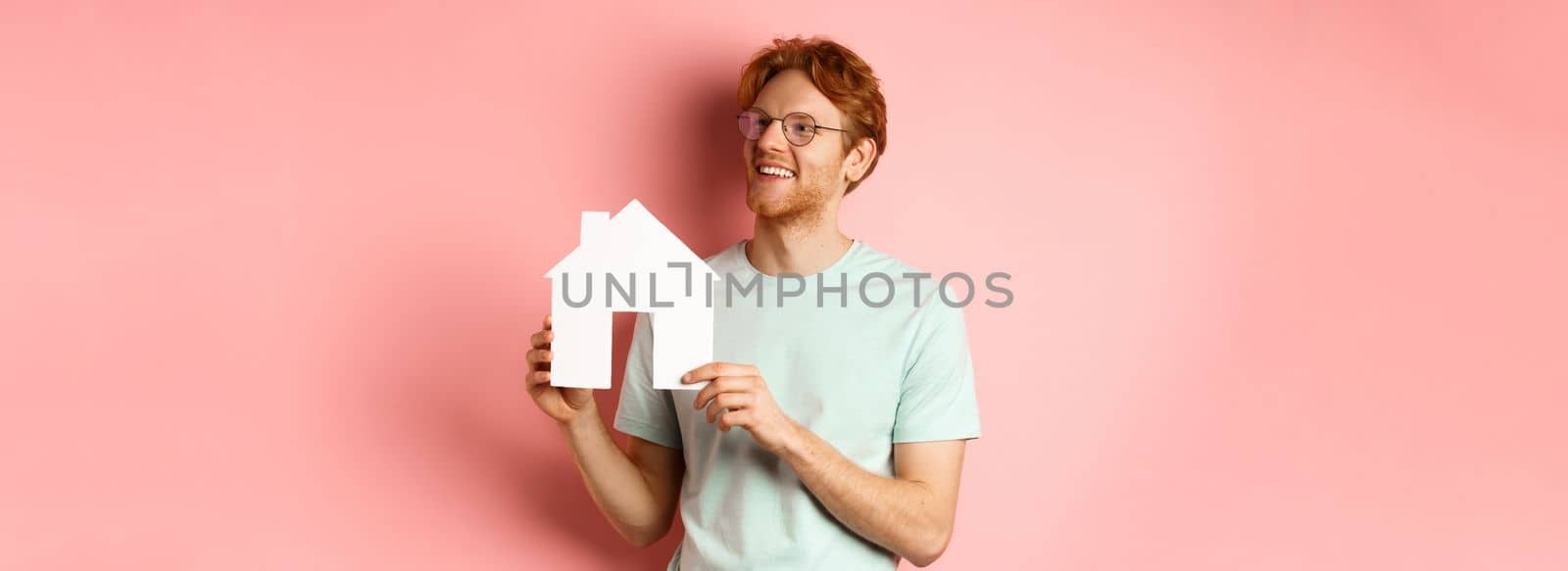 Real estate. Happy redhead man dreaming of buying property, looking right and showing paper house cutout, standing over pink background.