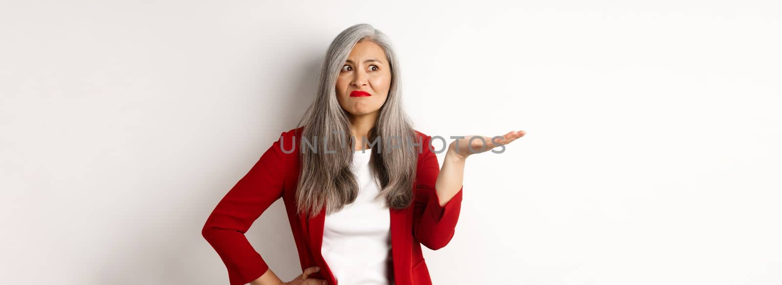 Annoyed and confused asian business woman spread hand sideways and shrugging, looking questioned left, white background.