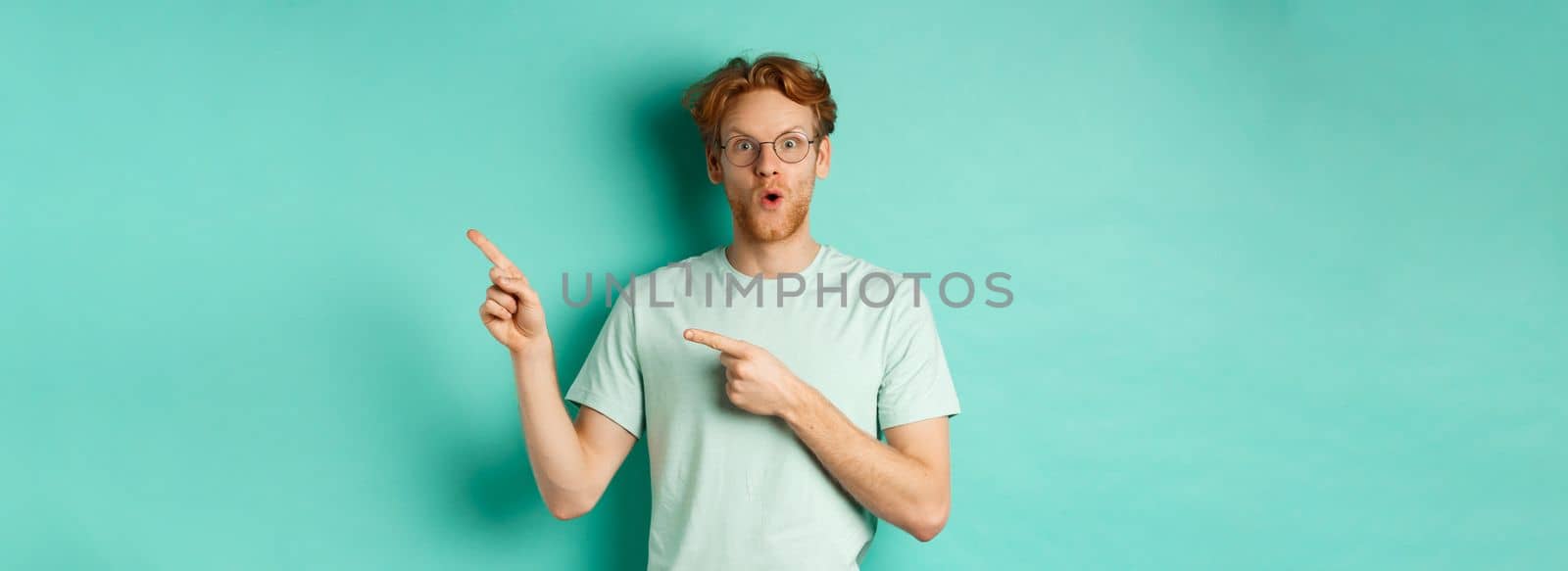 Surprised redhead guy in glasses and t-shirt checking out special deal, pointing at upper right corner promo and gasping in awe, showing banner, standing over turquoise background.