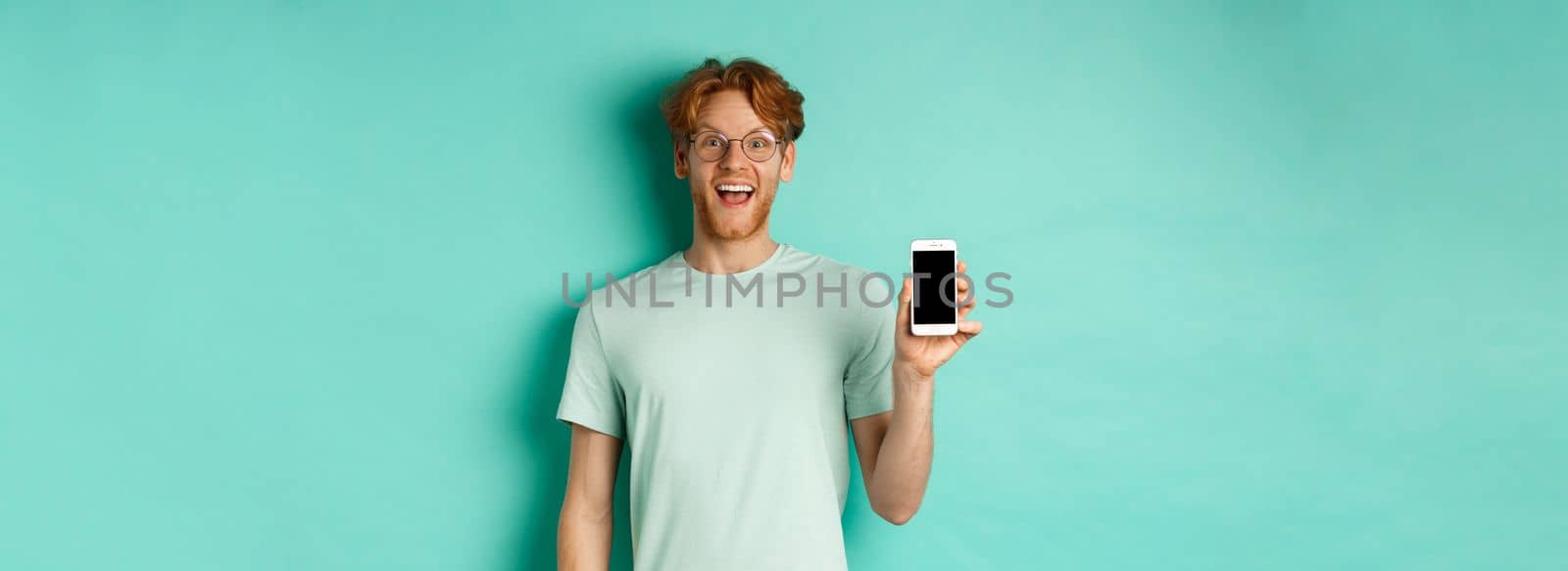 Online shopping. Cheerful redhead man in glasses and t-shirt showing smartphone screen and smiling, standing over mint background.