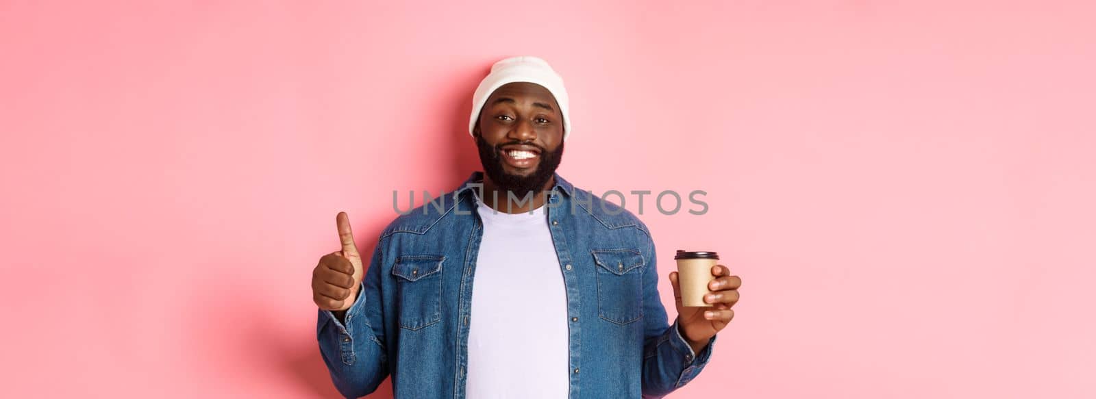 Handsome african-american hipster man showing thumb up, drinking coffee and recommending cafe, standing over pink background.