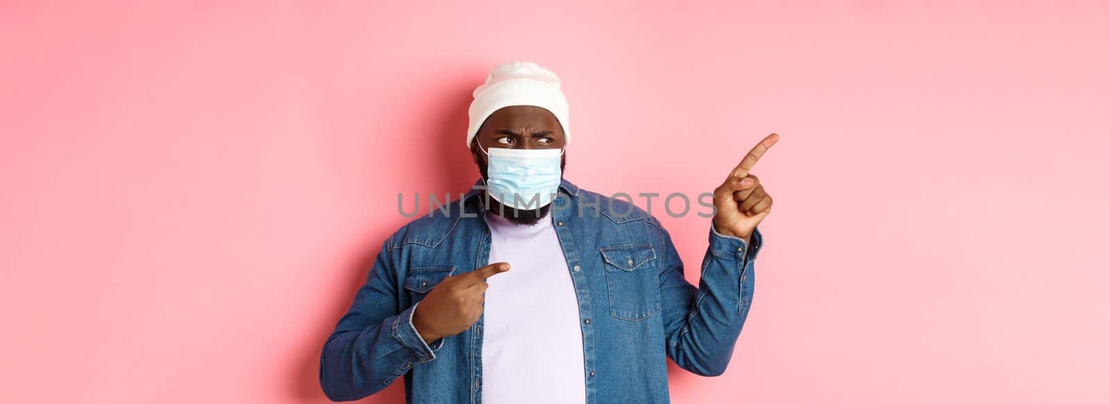 Coronavirus, lifestyle and global pandemic concept. Confused and disappointed african-american guy in face mask pointing fingers left, staring upset with camera, pink background by Benzoix