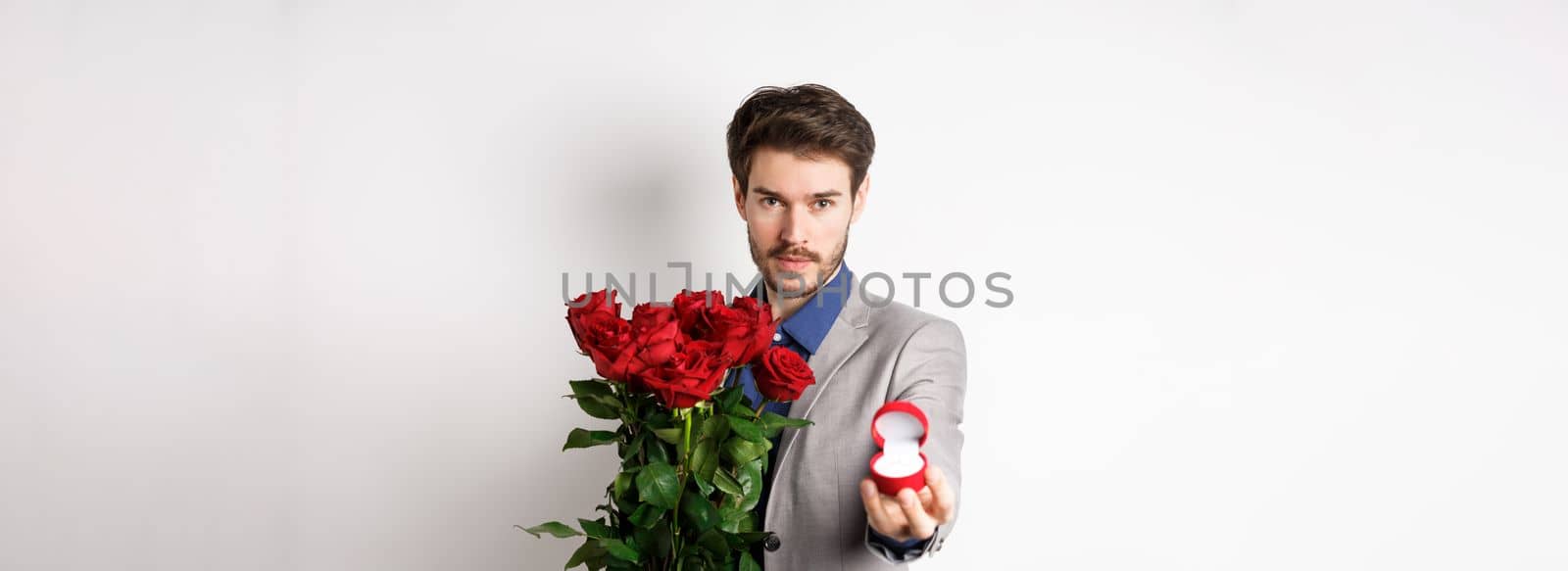 Handsome young man making a marriage proposal, stretch out hand with engagement ring and holding red roses, asking to marry him, looking confident at lover, white background.