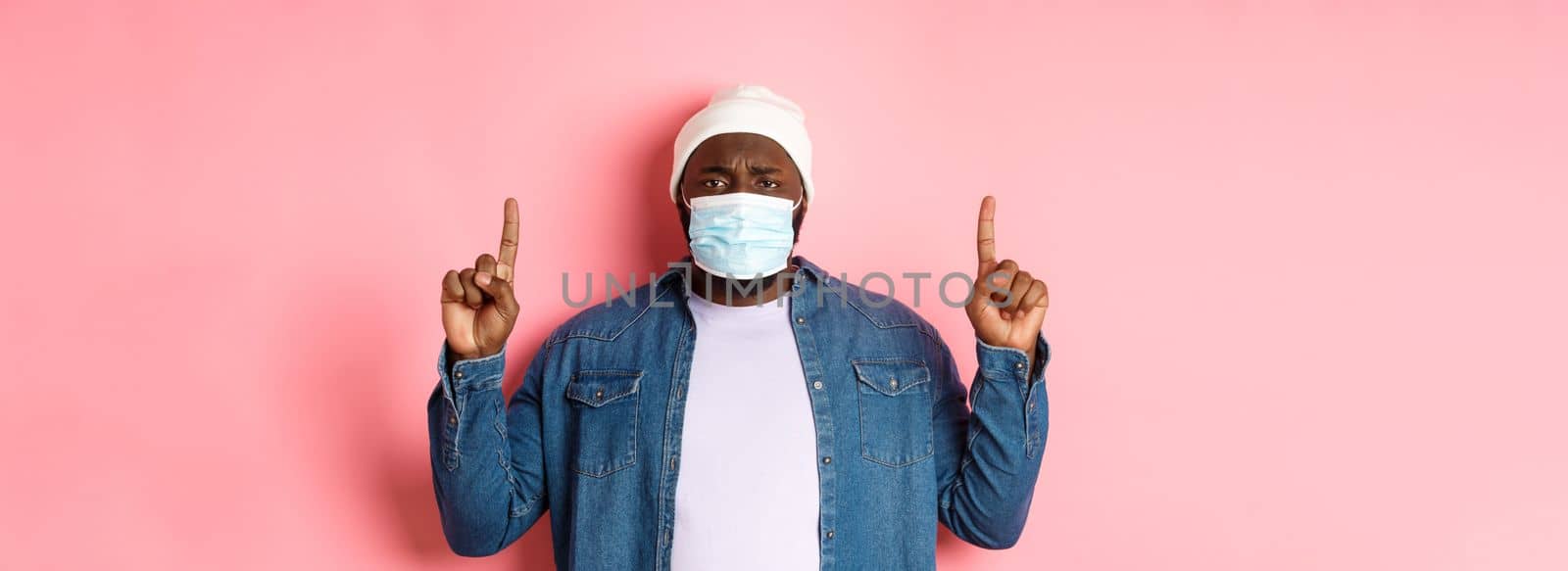 Coronavirus, lifestyle and global pandemic concept. Upset Black man in face mask frowning, pointing fingers up, showing advertisement, standing over pink background.