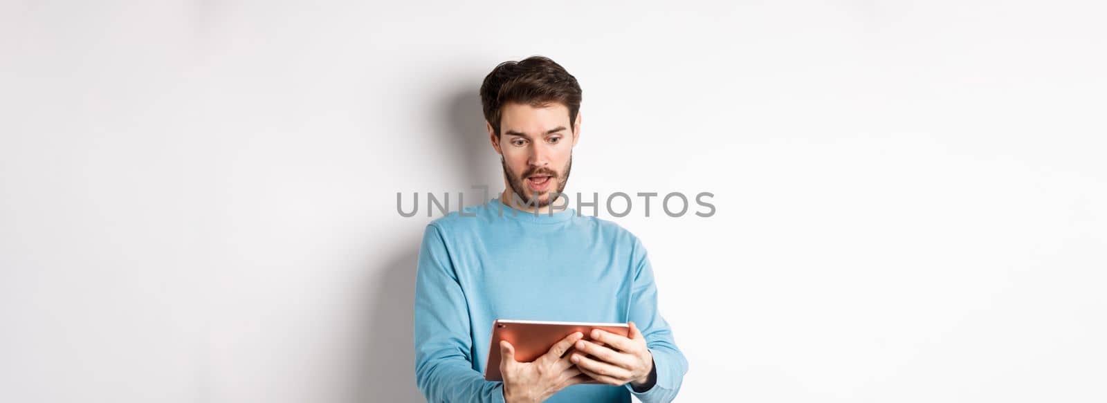Image of man looking curious at digital tablet, reading interesting news online, standing on white background. Copy space