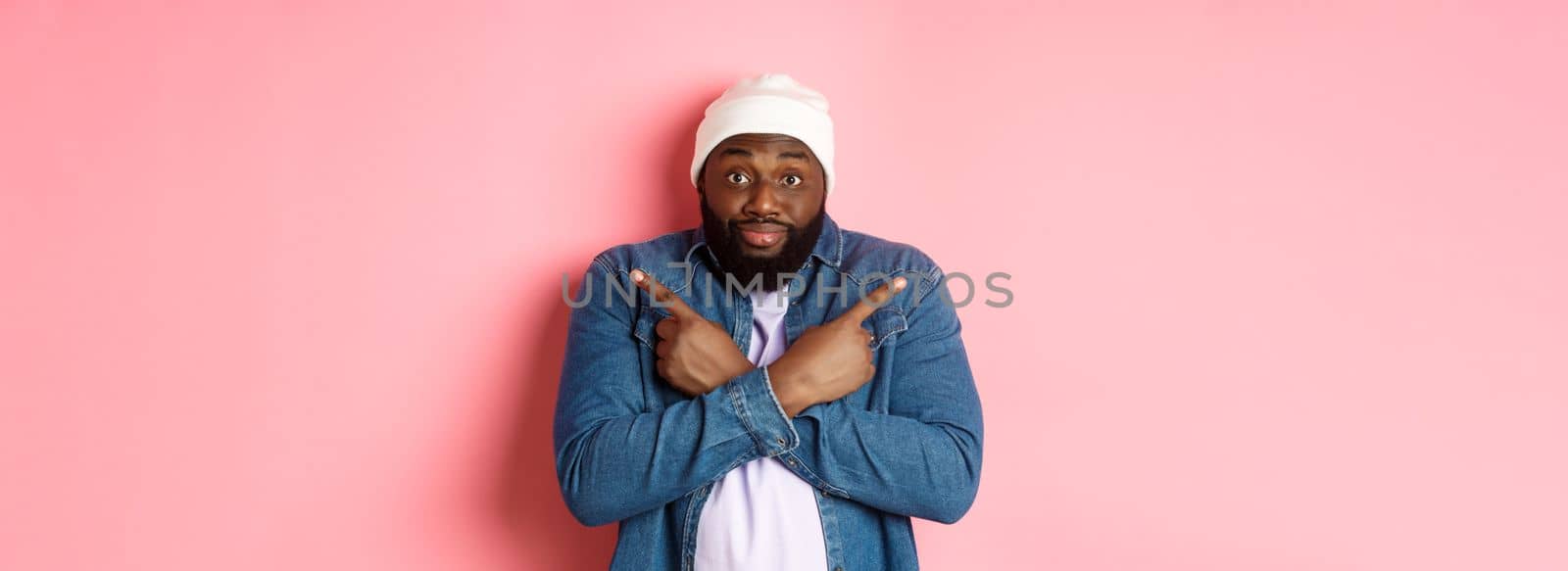 Confused Black man asking help with choice, pointing sideways and staring at camera, making decision, standing over pink background.