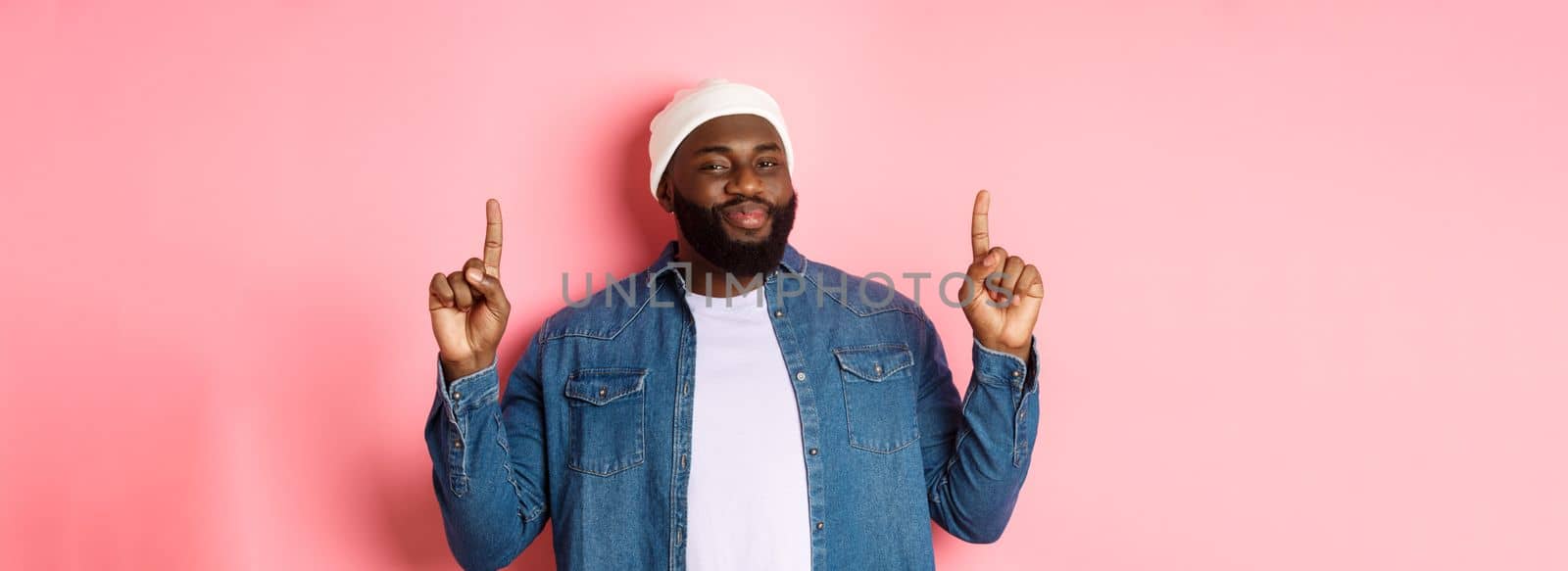 Pleased handsome Black man nodding in approval and checking out good promo, pointing fingers up at offer, showing best deal, pink background.