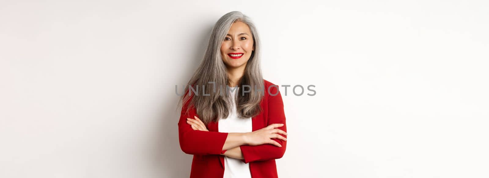 Business people. Smiling asian senior woman in red blazer, cross arms on chest and looking professional, standing over white background by Benzoix