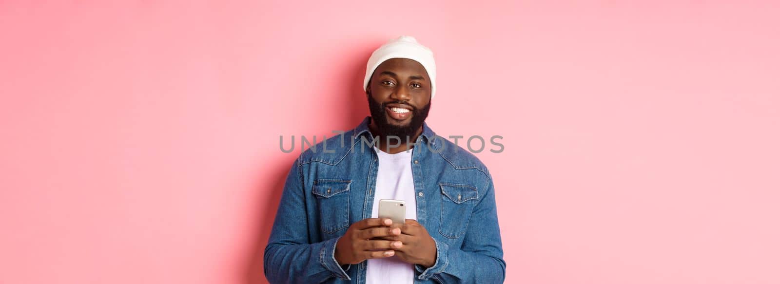 Handsome african-american guy using smartphone, smiling at camera, standing over pink background.