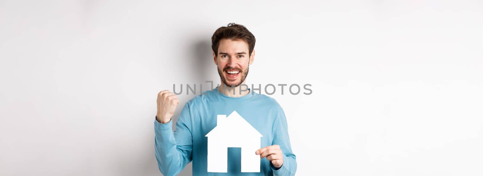 Real estate and insurance concept. Cheerful man buying property and celebrating, saying yes and showing paper house cutout, standing on white background.